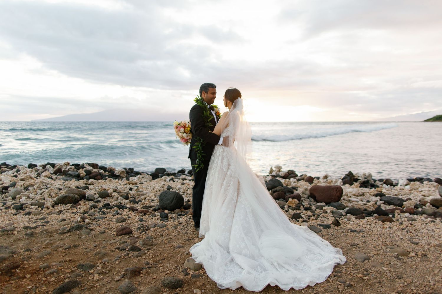 A couple in traditional Hawaiian leis poses on a rocky beach for their wedding portraits.
