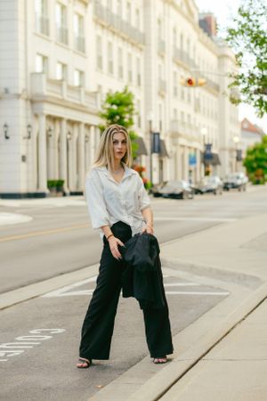Woman in white button-down shirt and black wide-leg pants poses on city street with classic architecture in background.