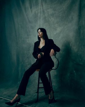 Woman in black suit seated on stool against moody gray backdrop in elegant studio portrait.