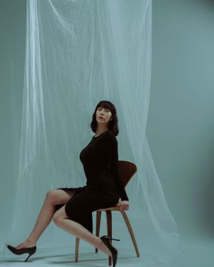 Stylish figure in black dress poses on wooden chair against ethereal white draped fabric backdrop in moody studio setting.