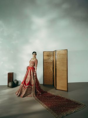 Ornate red and gold bridal lehenga dress displayed against wooden panels in a minimalist studio setting.