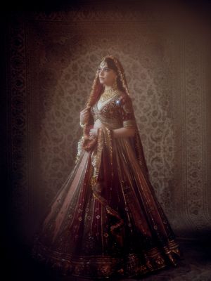 Dramatic low-light portrait of an ornate red and gold Indian wedding lehenga against a patterned background.