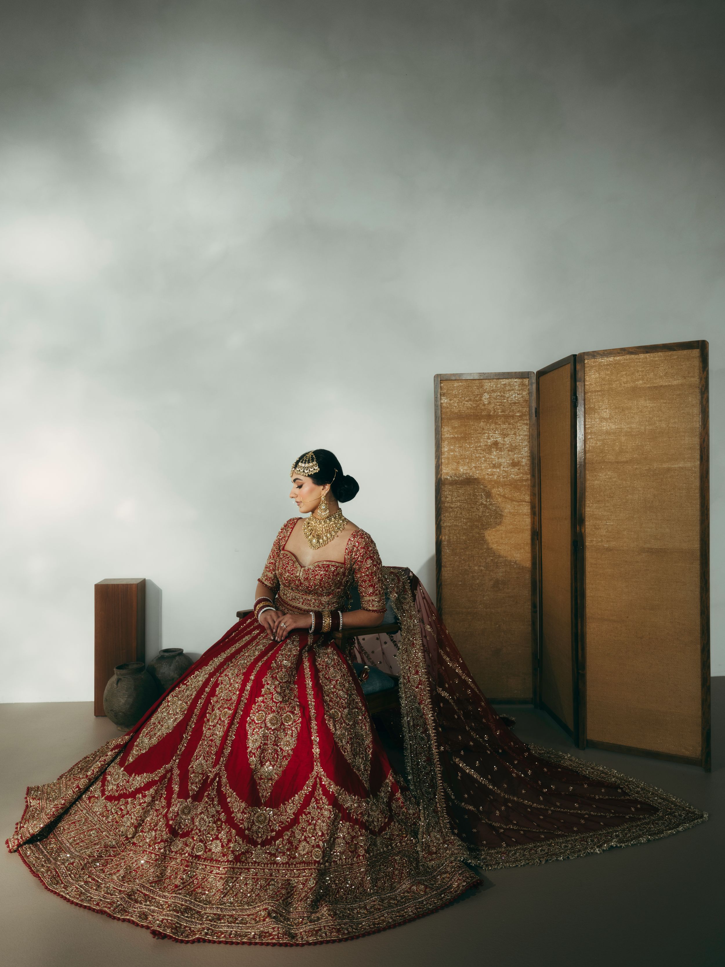 A person wearing an ornate red and gold traditional Indian bridal lehenga poses elegantly against a minimalist backdrop.