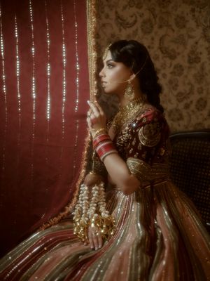 A person in traditional South Asian bridal attire with gold jewelry and embroidered clothing poses against a patterned wall.
