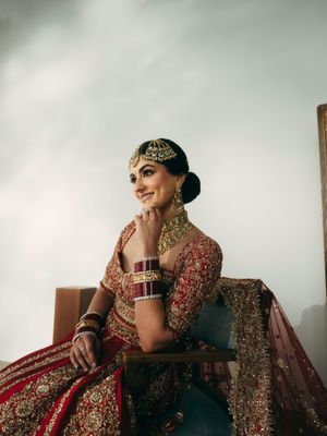 A bride in a traditional red and gold embellished Indian wedding lehenga poses elegantly on a wooden chair.
