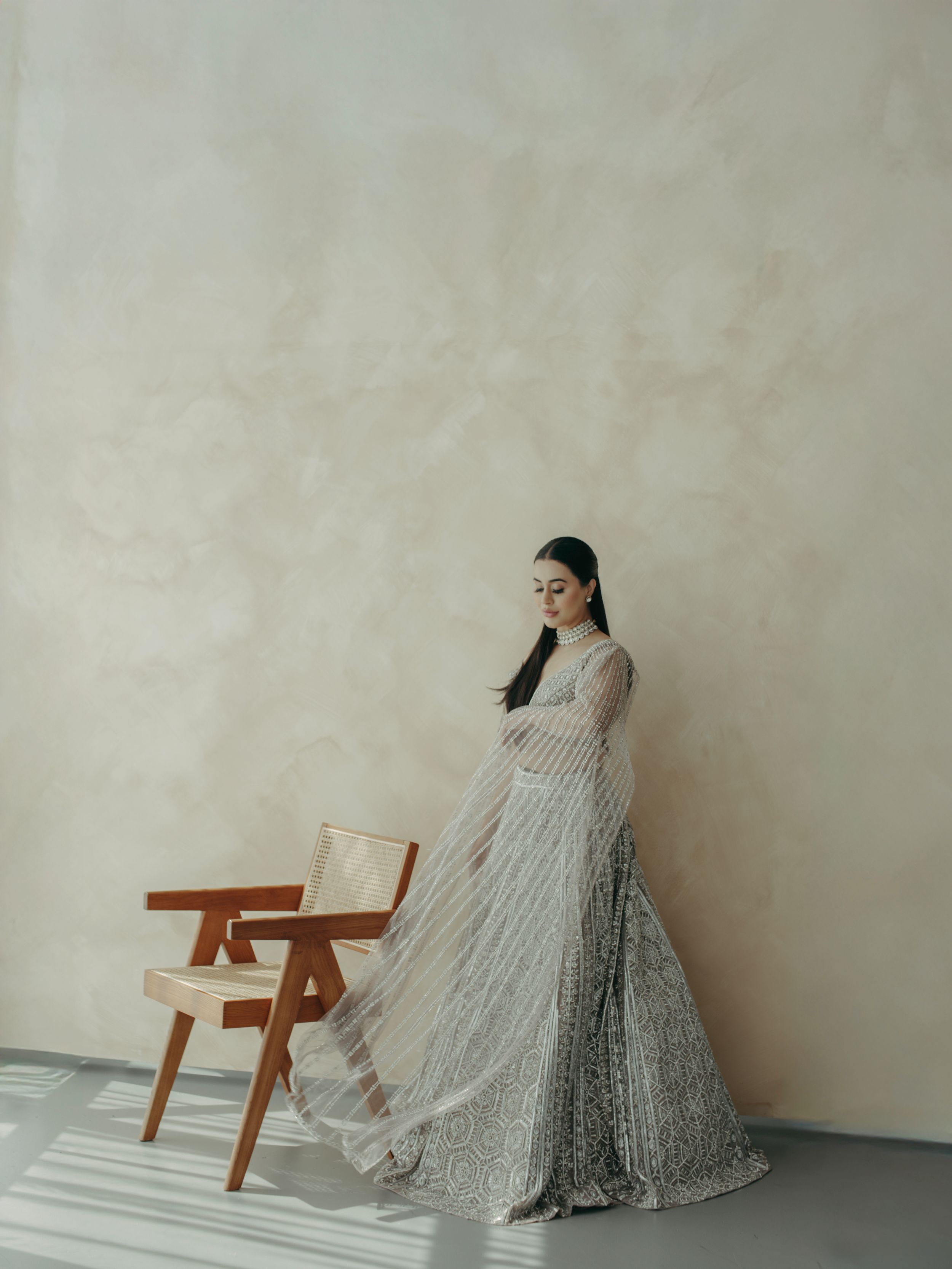 A woman in a silver sequined floor-length gown poses elegantly next to a wooden chair against a minimalist beige wall.