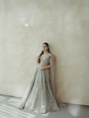 A person wears an elegant silver-gray embellished lehenga gown against a minimalist beige wall with soft lighting.
