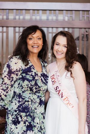 Mother and daughter smile at the camera. The daughter is wearing white and celebrating her bridal shower.