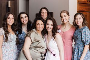 A group of seven women smiling at the camera in dresses. A woman in a white dress celebrates her bridal shower.