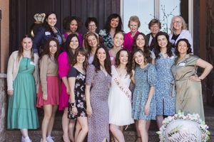 A group of nineteen women of various ages are smiling at the camera in dresses. A woman in a white dress celebrates her bridal shower.