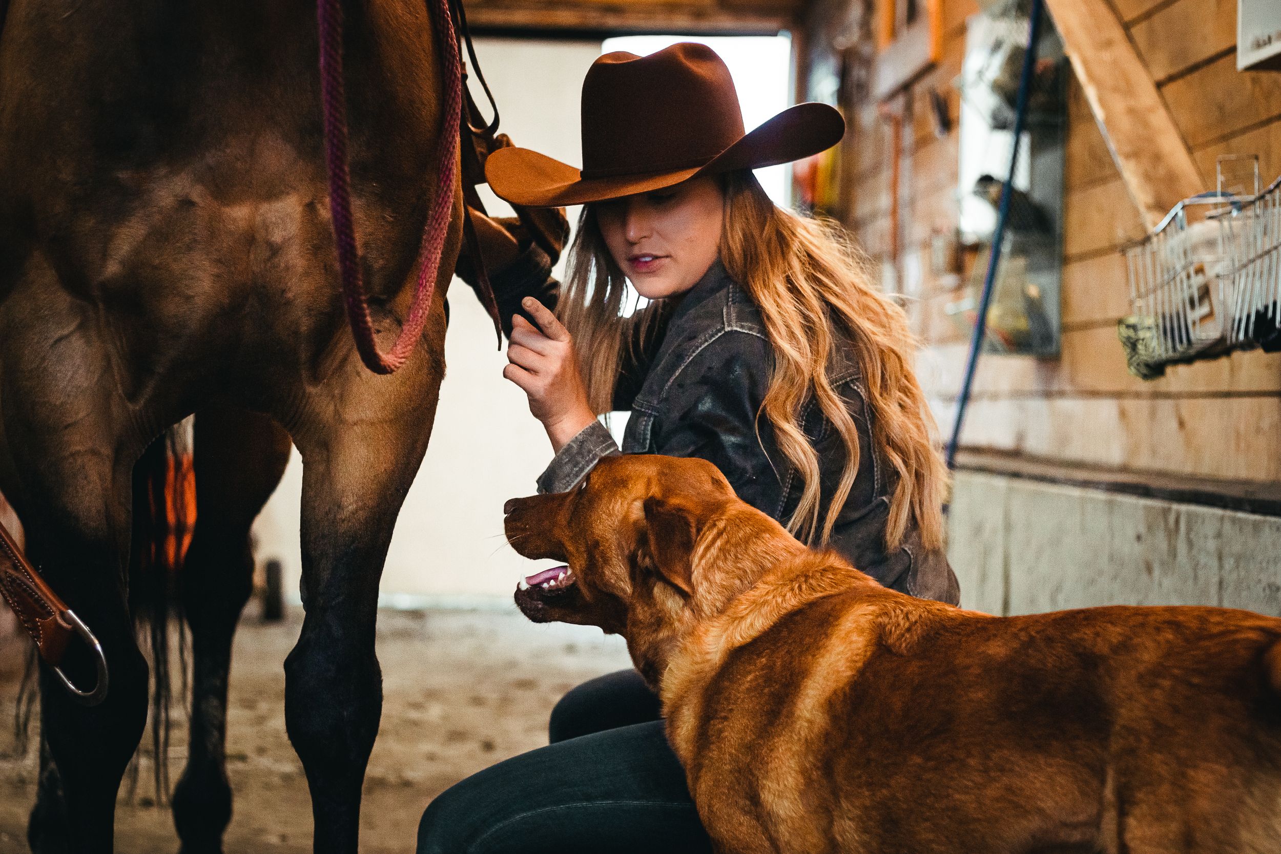 A western styled woman looks at her dog while crouched beside her horse.
