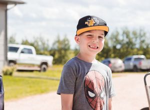 A young boy smiles and winks at the camera. He wears a Spiderman shirt and black ball cap. Cars in the background.