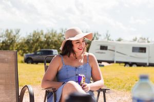 A woman sits laughing at someone while wearing a blue dress, tan straw hat, and holding a drink.