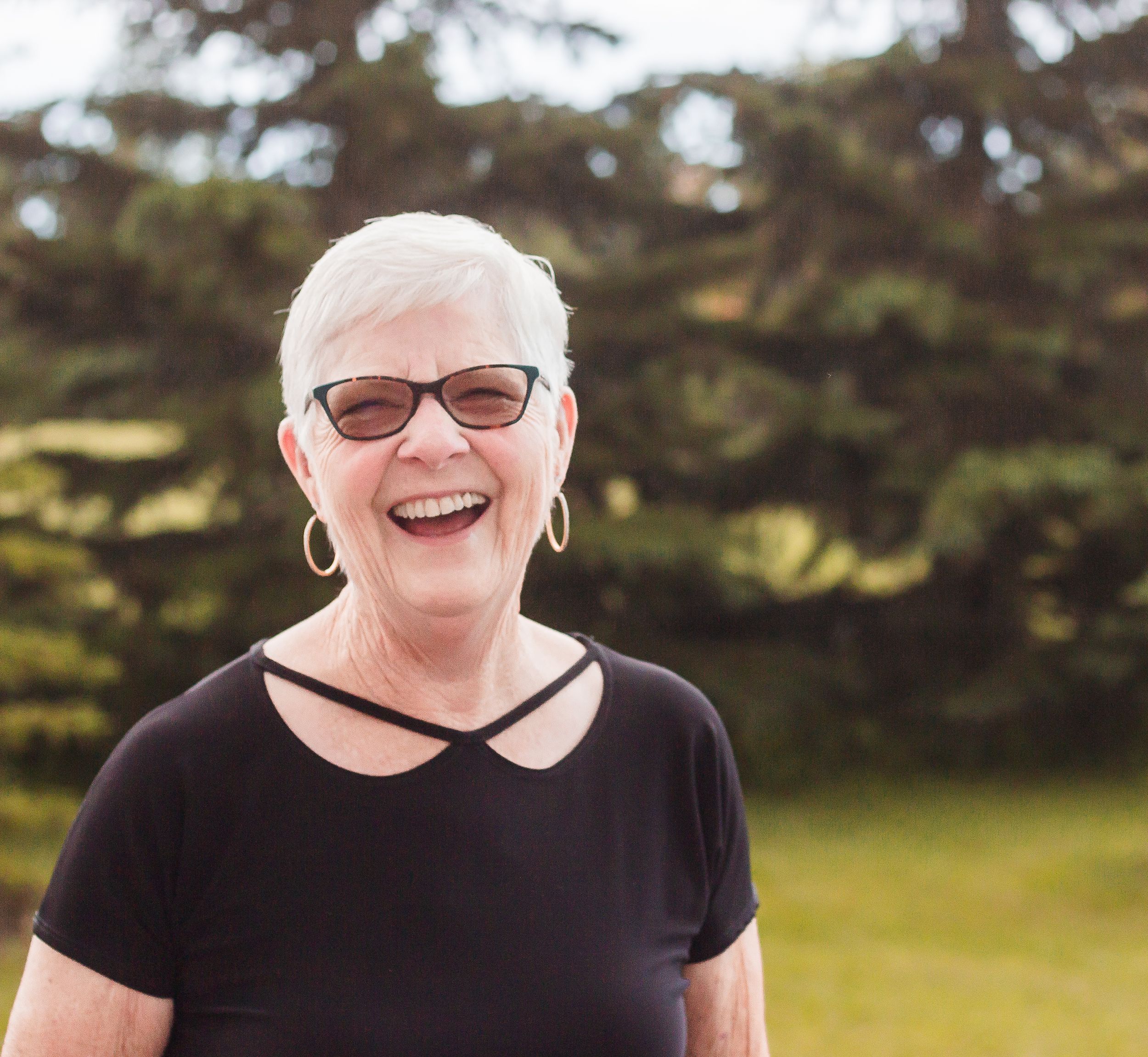 An older woman laughs towards the camera. She wears sunglasses, hooped earrings, and a black shirt.