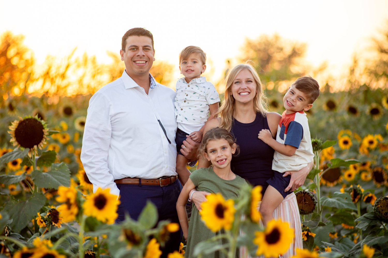 Happy family of five smiles together in a vibrant sunflower field at golden hour.