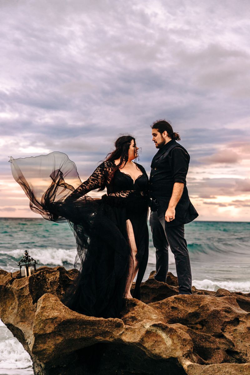 Creative couples' anniversary on the beach Shes wearing black lace gown that's blowing in the wind at sunset by Photogenic Fox Photography