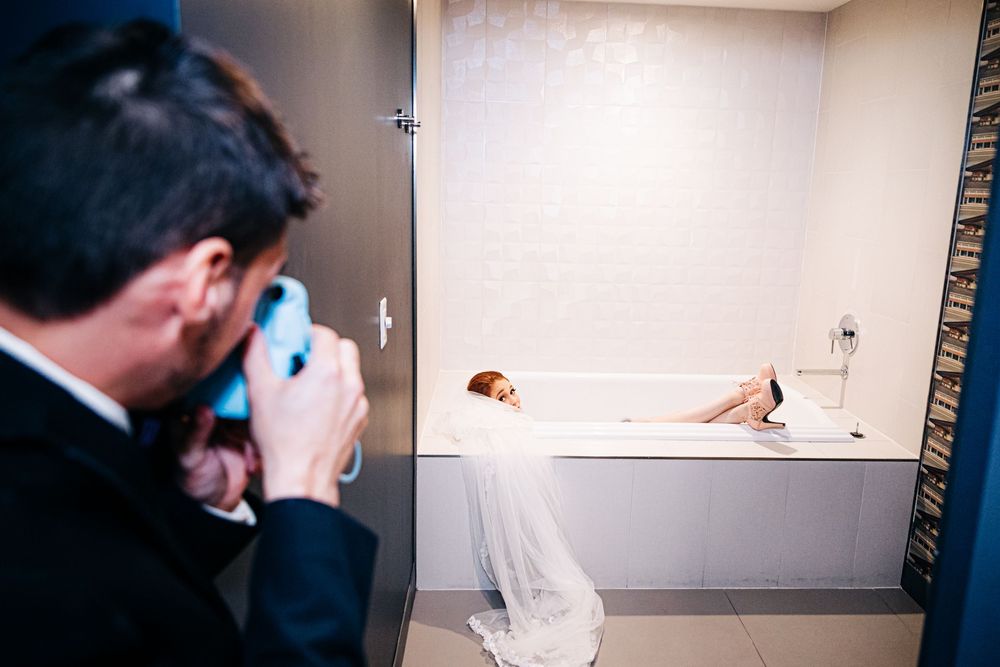 Wedding Groom using Polaroid camera to take picture of bride being cute in the hotel bathtub by Photogenic Fox Photography