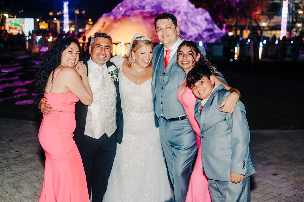 A wedding party poses together at night with colorful lighting in the background is sandy tree at West Palm Beach Lake Pavilion