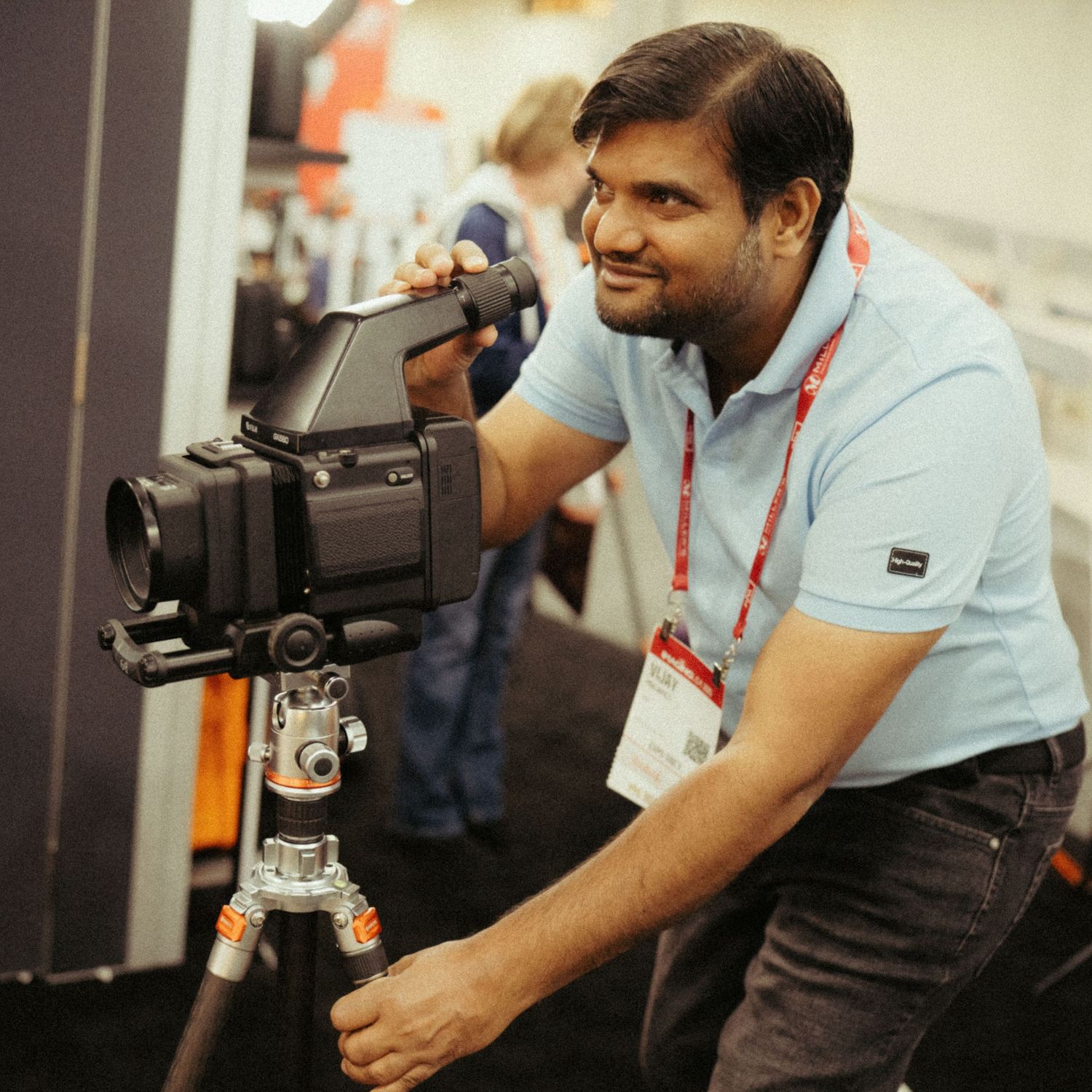 Camera operator wearing light blue polo shirt adjusts professional video camera mounted on tripod at industry event.
