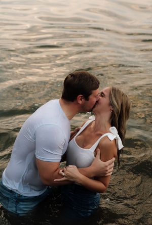 A couple shares a romantic kiss while standing in shallow water, both wearing white tops and jeans.