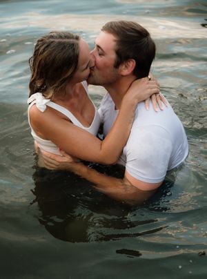 Couple sharing a romantic kiss while standing in water, both wearing white wet clothing in an outdoor setting.