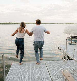 Couple holding hands running off a dock into a lake, wearing jeans and casual tops on a cloudy day.