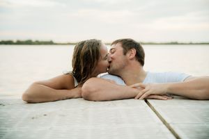 Couple sharing a romantic kiss while lying on a wooden dock overlooking a calm lake on a cloudy day.