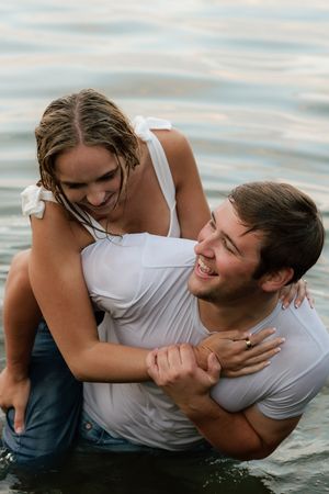 Happy couple laughing and embracing in water, wearing wet white shirts during a romantic outdoor photo session.