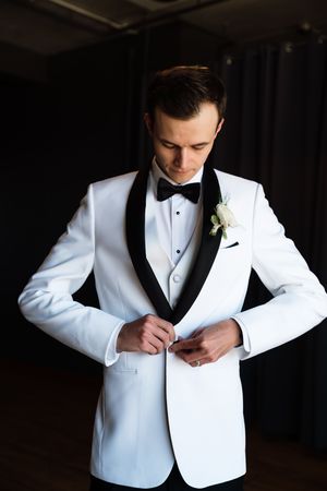 A groom in a white tuxedo with black lapels and bow tie buttons his jacket, wearing a white rose boutonniere.