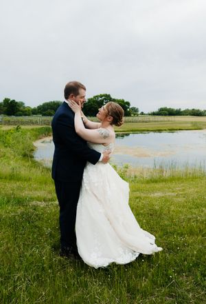 A bride and groom embrace romantically in a green field beside a pond on their wedding day.