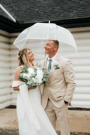 Bride and groom smiling under a clear umbrella on their wedding day, bride holding white floral bouquet.