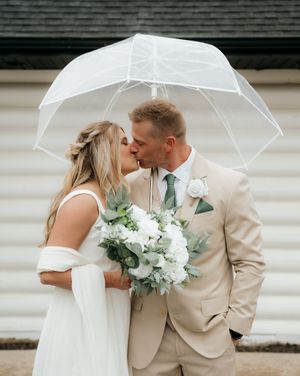 Bride and groom kissing under a clear umbrella, bride holding white floral bouquet, groom in beige suit with green tie.