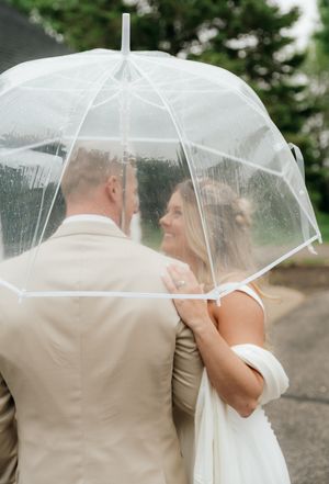 A bride and groom share a romantic moment under a clear dome umbrella in the rain on their wedding day.