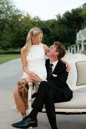 A bride in a short white dress and groom in a black suit share a romantic moment on an elegant white sofa outdoors.