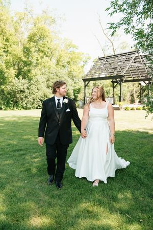 Bride and groom walking hand in hand on green grass near a wooden pergola.