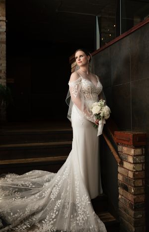 Bride in lace wedding gown with train holds white bouquet while posing on staircase.