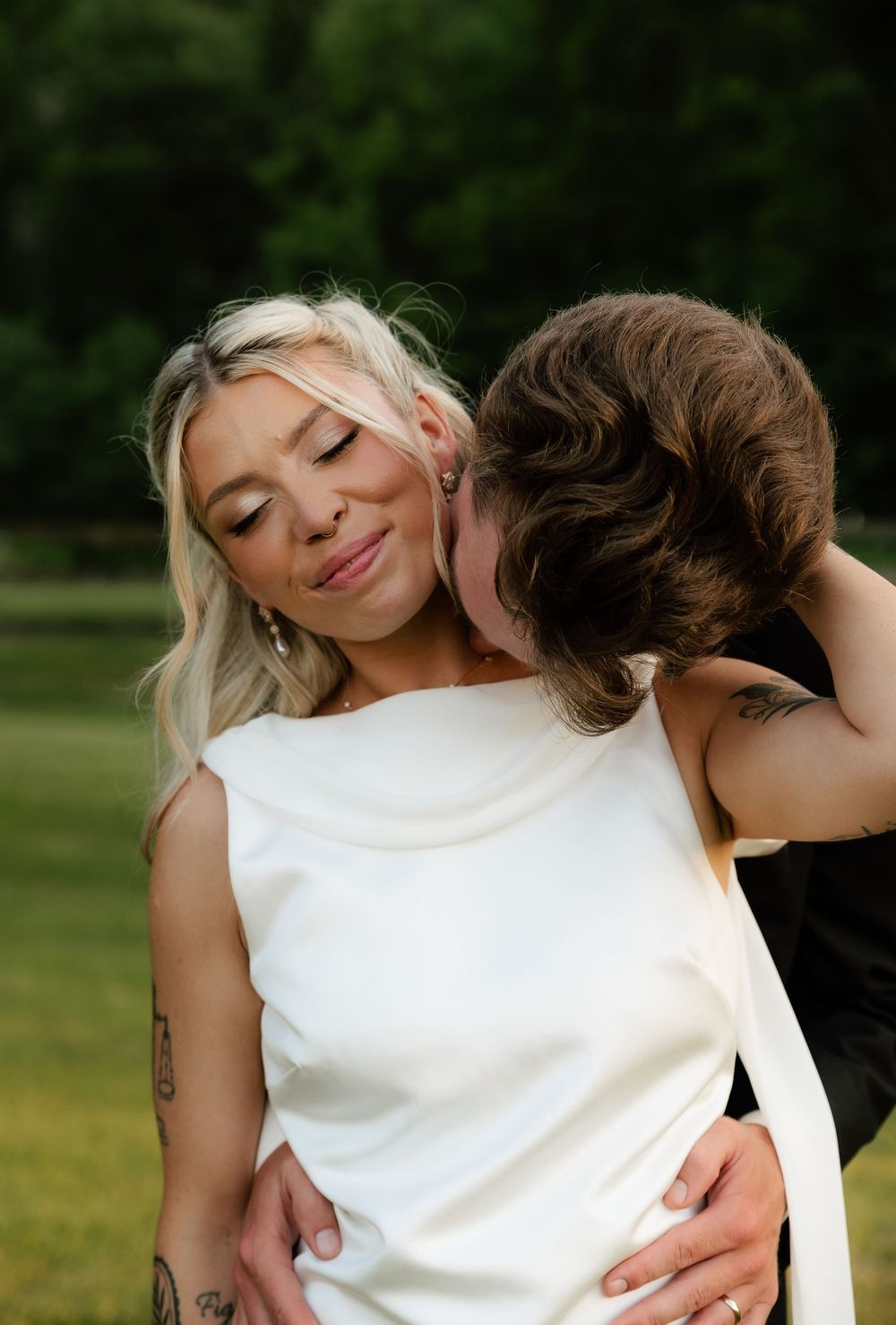 A bride in a white gown smiles as her partner kisses her neck outdoors in a lush green setting.