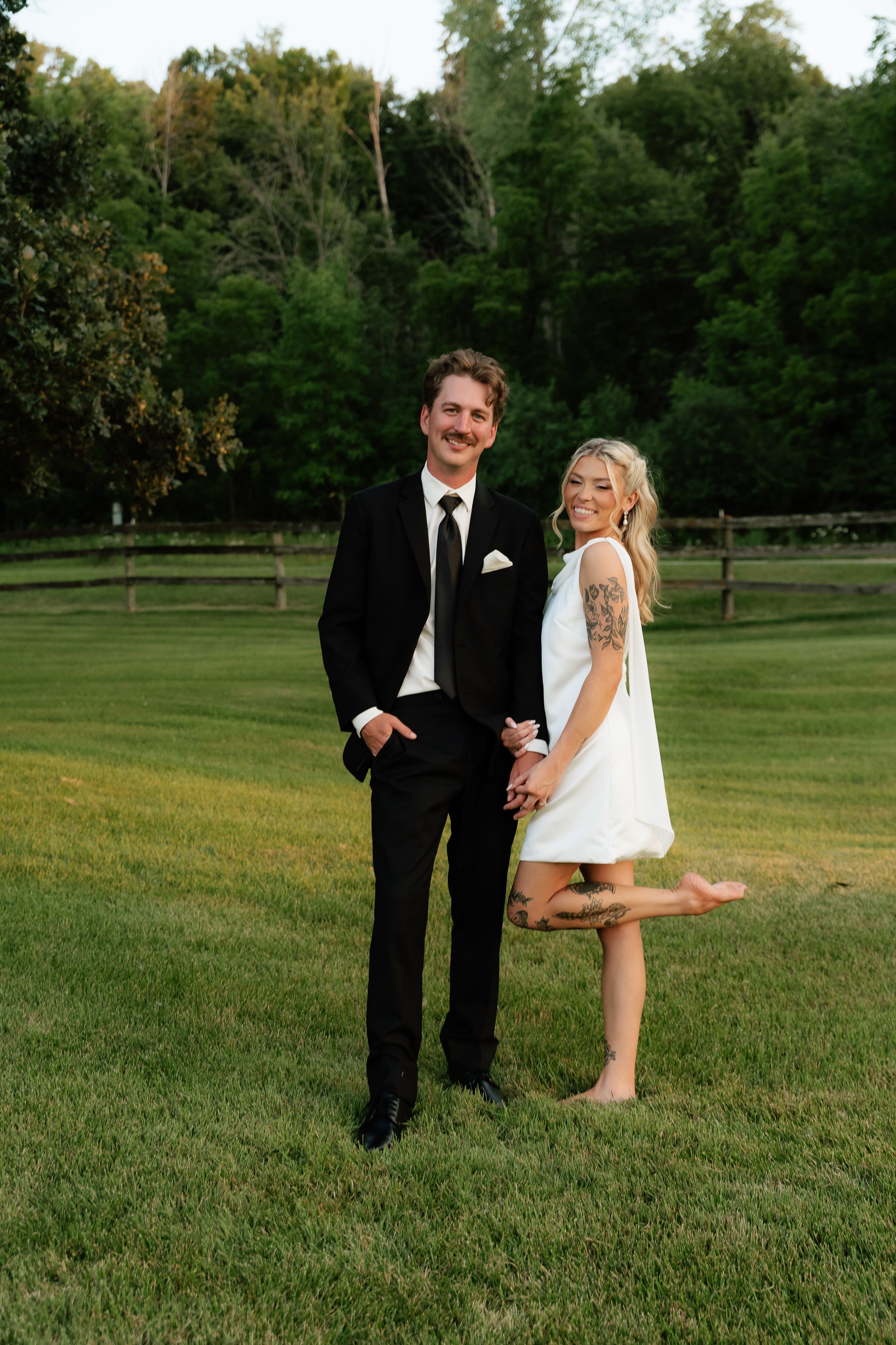 A couple poses on green grass, man in black suit, woman in white mini dress with tattoo, trees in background.