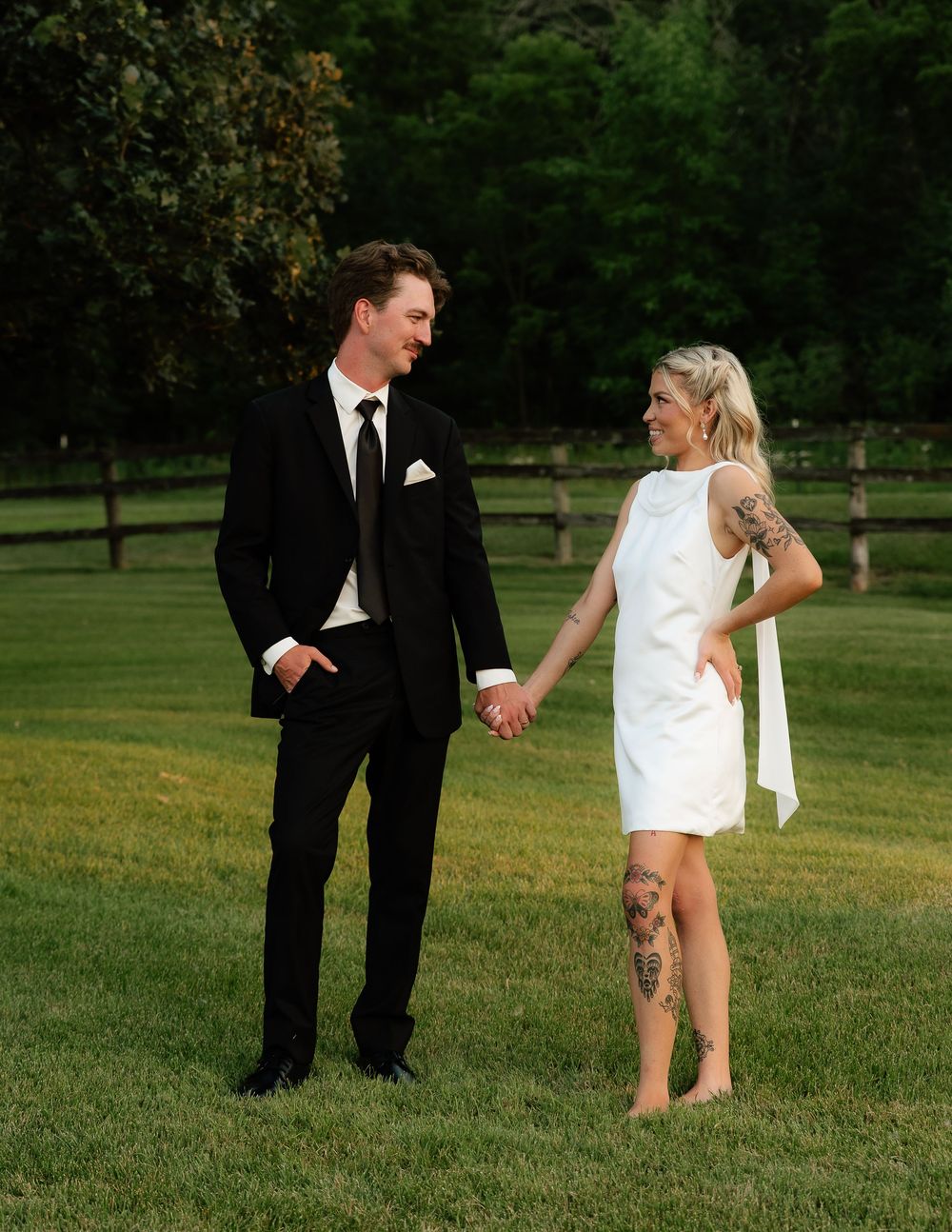 Couple holding hands on green lawn, man in black suit, woman in white mini dress with leg tattoo, outdoor wedding photo.