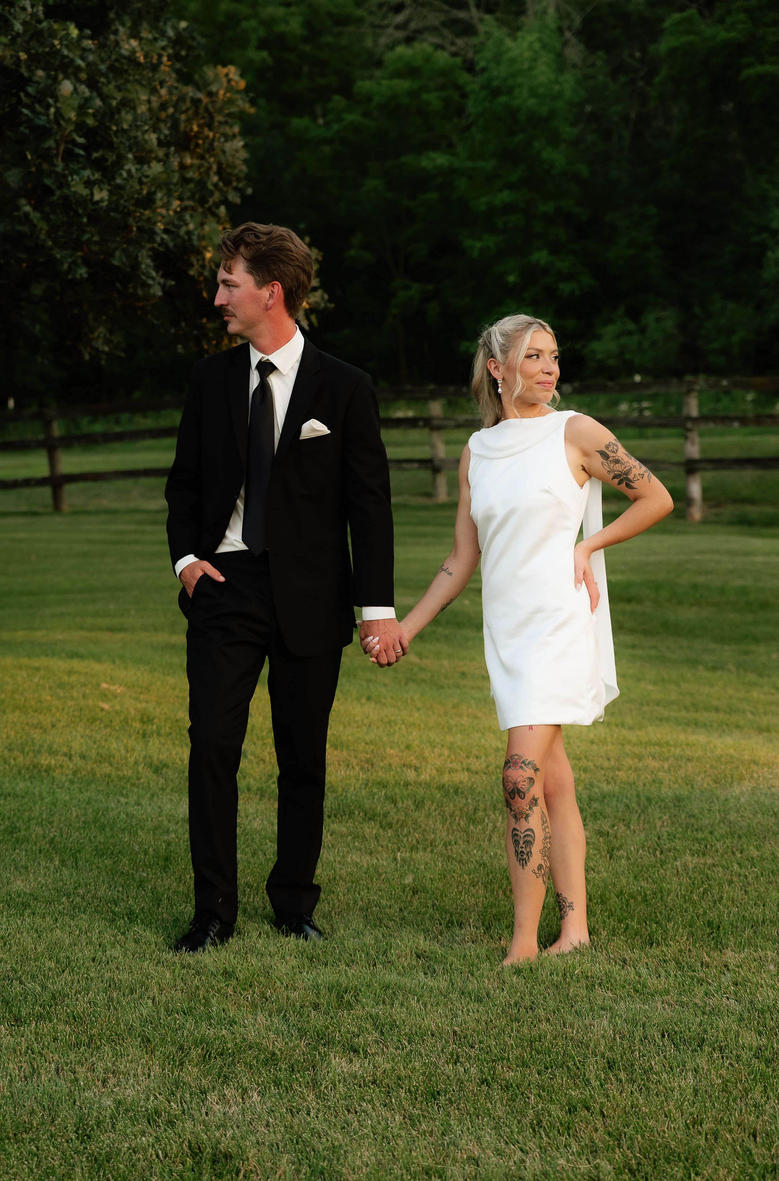Couple in wedding attire holding hands on green lawn, groom in black suit, bride in white mini dress with leg tattoos.