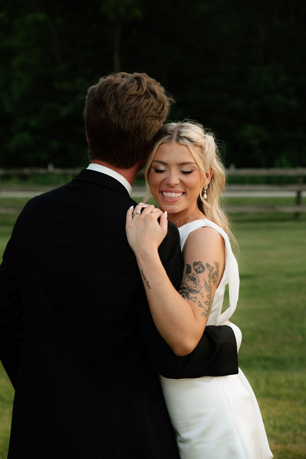 Smiling bride with tattoos in white gown embraces groom in black suit outdoors at golden hour wedding celebration.