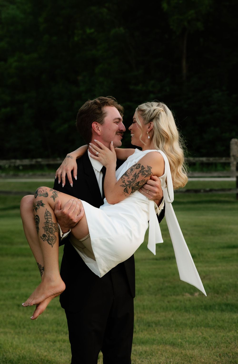 Groom in black suit carries bride in white dress outdoors on green lawn, both smiling and sharing a romantic moment.
