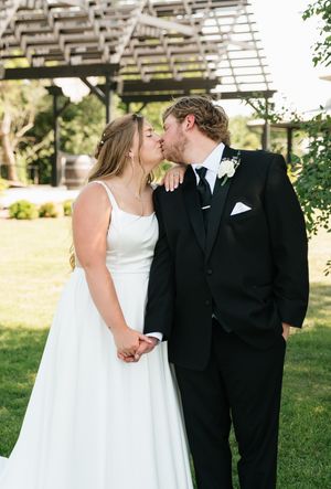 Bride and groom share a kiss while holding hands on green lawn with rustic pergola backdrop.