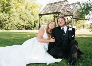 Smiling bride in white gown and groom in black tuxedo sit together on lush green grass outdoors.