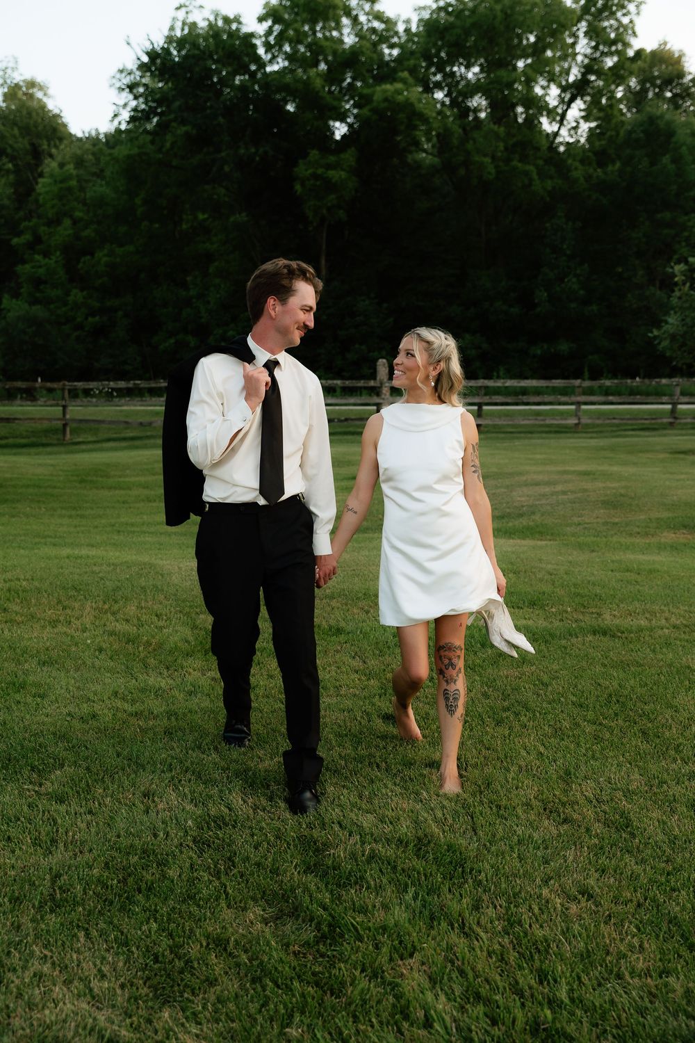 A bride and groom walk hand-in-hand on green grass, she holds heels barefoot, he carries his jacket over his shoulder.