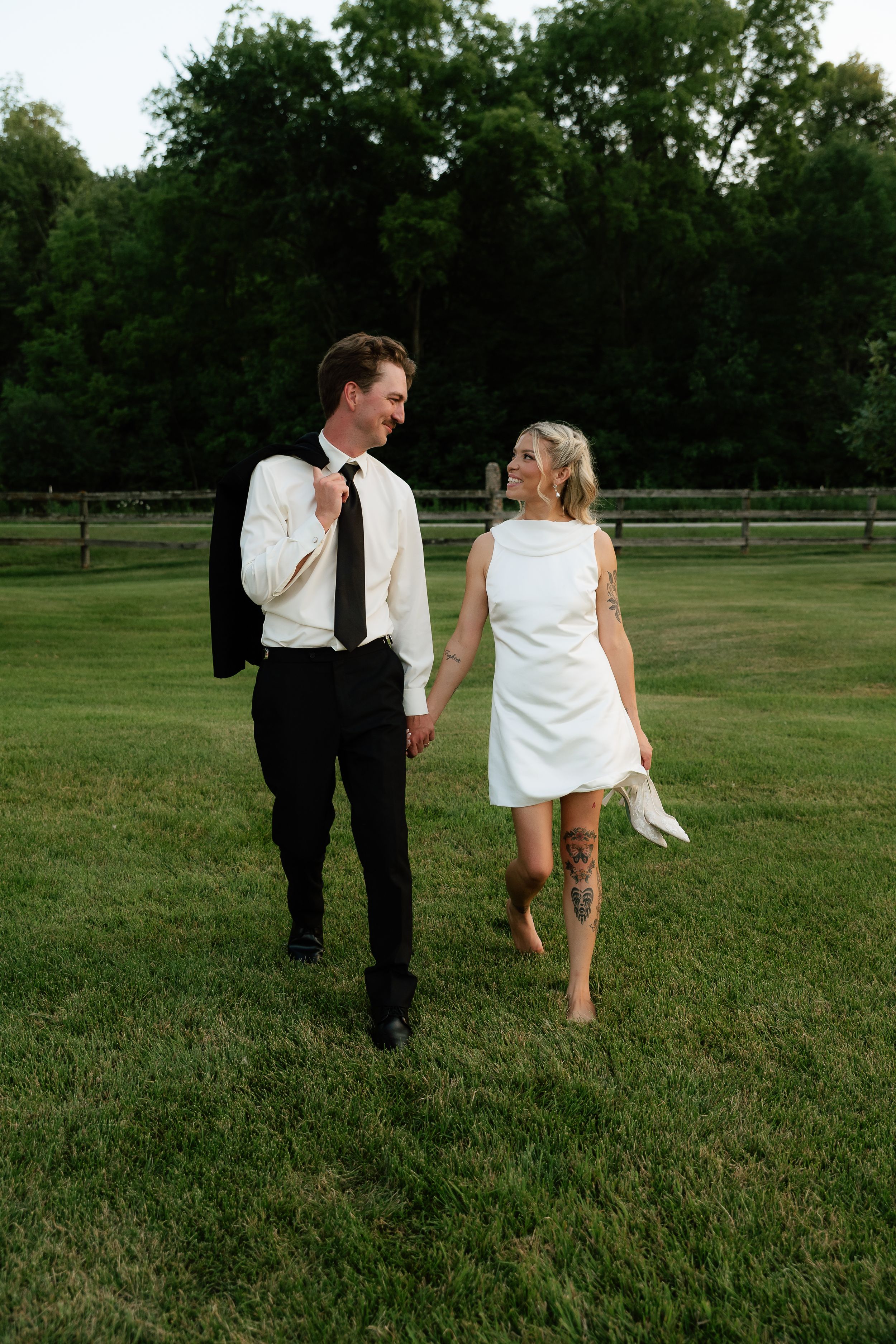 A bride and groom walk hand-in-hand on green grass, she holds heels barefoot, he carries his jacket over his shoulder.