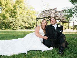 Bride and groom sitting together on green grass in front of a wooden pergola on their wedding day.