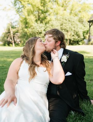 Bride and groom share a romantic kiss while sitting on green grass outdoors, wearing wedding attire on their special day.