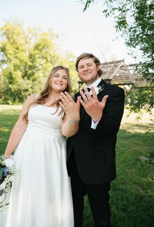 Newlywed couple in wedding attire smiling and showing off their wedding rings outdoors in a lush green garden setting.