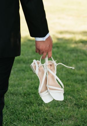 A groom in a black suit holds white strappy bridal heels over green grass at a wedding.